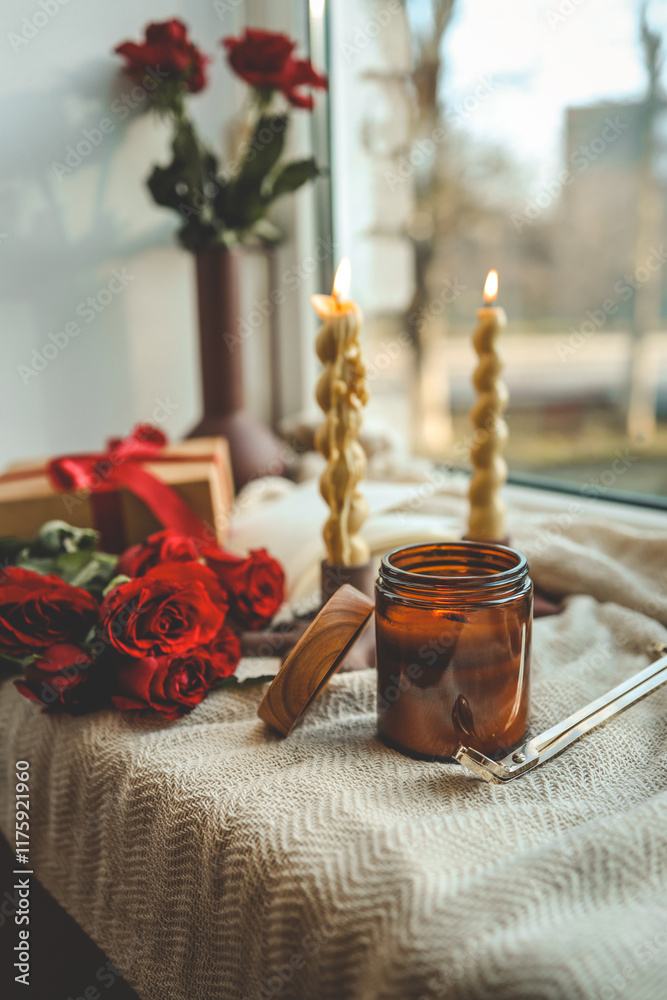Two twisted candles burning in modern holders by a windowsill, accompanied by a red ribbon gift box, red roses, and a vase with roses, creating a cozy ambiance with natural light Two twisted candles burning in modern holders by a windowsill, accompanied by a red ribbon gift box, red roses, and a vase with roses, creating a cozy ambiance with natural light