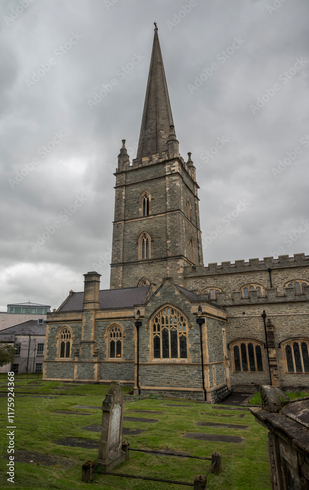 Obraz premium Saint Columb's Cathedral with a Gothic spire and weathered gravestones under an overcast sky, Londonderry, Northern Ireland