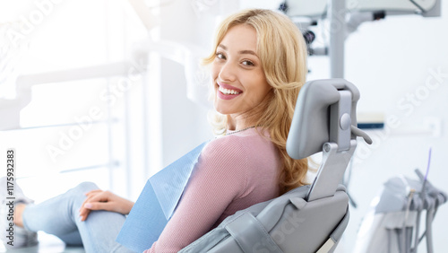 Smiling and relaxed young woman sitting at dental chair in modern clinic, waiting for her dentist. Happy attractive blonde lady attending luxury dental clinic, bacl view, copy space