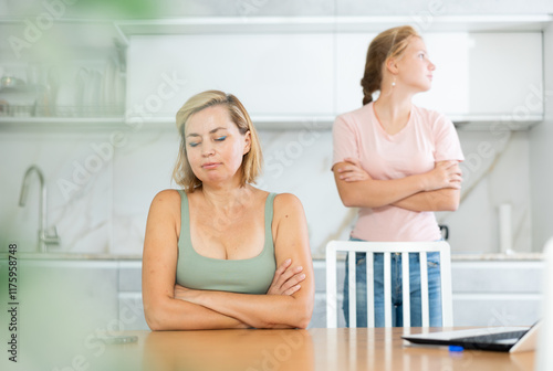 Portrait of disillusioned woman facing relationship problems with teenage daughter standing behind in blurred background in home kitchen photo