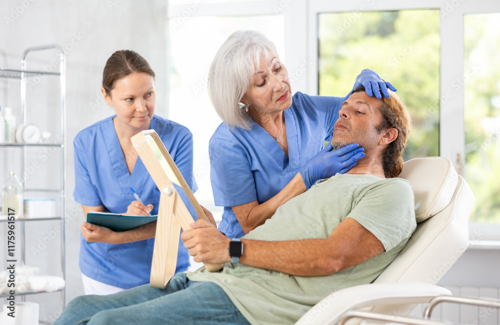Obraz premium Interested adult man looking at mirror while skilled female cosmetologist showing results of facial procedure in medical esthetic office. Female assistant filling out patient medical record