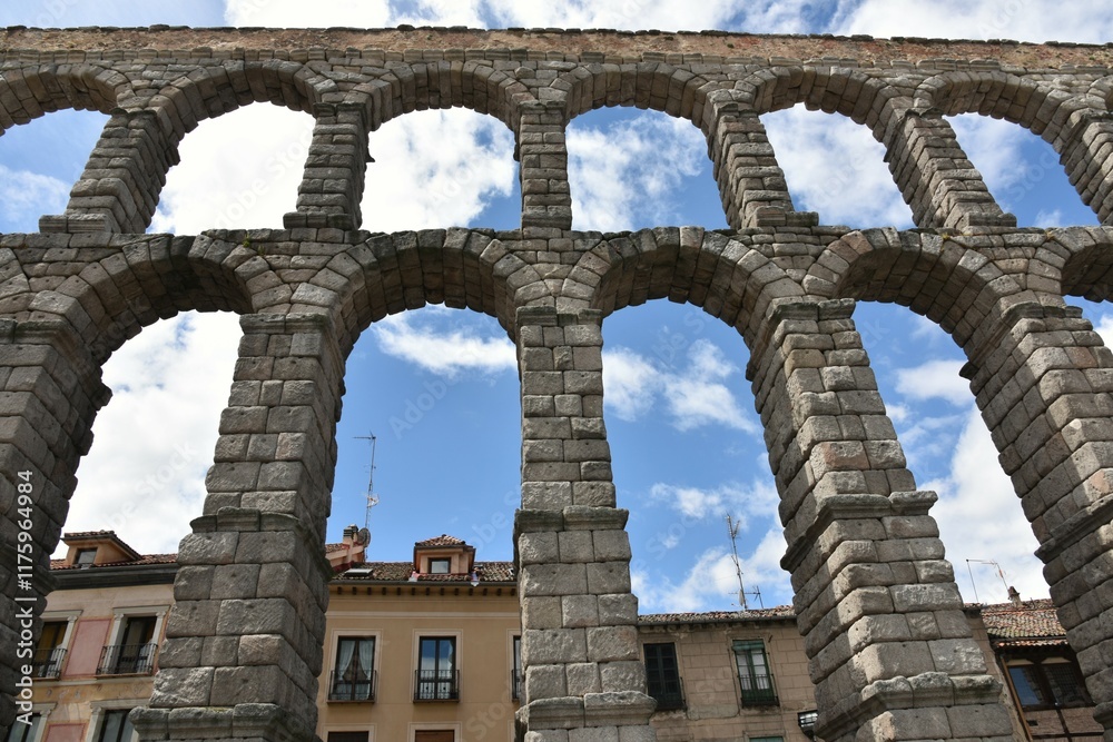 Central symmetric view of the arches of famous Roman Aqueduct of Segovia, Spain, in grey granite ashlar stone against a backdrop of contemporary houses and a slightly cloudy blue sky Central symmetric view of the arches of famous Roman Aqueduct of Segovia, Spain, in grey granite ashlar stone against a backdrop of contemporary houses and a slightly cloudy blue sky
