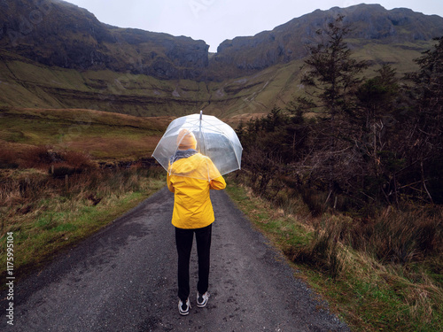 Travel and tourism in Ireland. Teenager girl with umbrella on a road and looking at majestic nature scenery of Gleniff Horseshoe drive with tall mountains. Model wearing yellow jacket and hat. photo