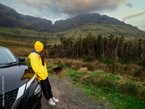 Travel and tourism in Ireland. Young woman standing by black car and looking at looking at majestic nature scenery of Gleniff Horseshoe drive with tall mountains. Model wearing yellow jacket and hat. photo