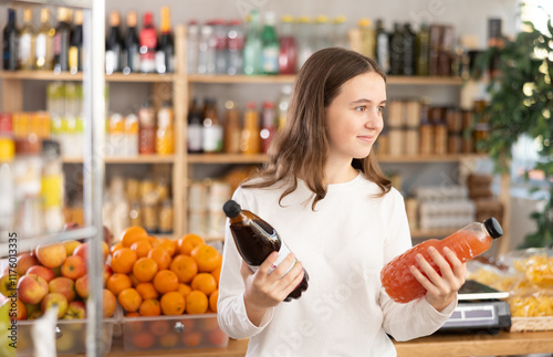 European teenage girl took various varieties of juices from shelf, chooses fruit drink for dinner with friends. Client buys drinks and snacks for buffet photo