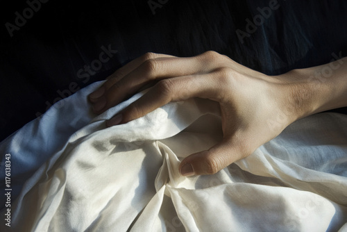 Depiction of a hand gently resting on a soft, draped fabric in a dimly lit environment highlighting the contours and textures of the skin and cloth