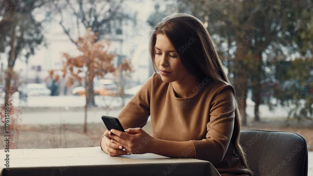A young woman sits at a table, intently using her smartphone. She has long hair and a neutral expression as she observes the screen, with blurred outdoor scenery in the background. A young woman sits at a table, intently using her smartphone. She has long hair and a neutral expression as she observes the screen, with blurred outdoor scenery in the background.