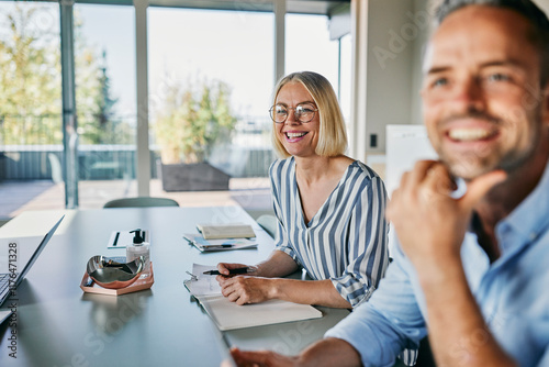 Smiling Colleagues Engaged in a Productive Meeting at a Modern Office. The colleagues are sitting next to each other, notebooks in front of them. They look up, smiling, at another colleague.