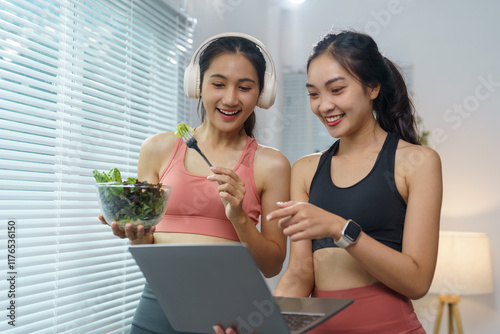 Two young, sporty asian women enjoying healthy salads while using a laptop at home, discussing their online fitness training program and sharing tips for a balanced lifestyle photo