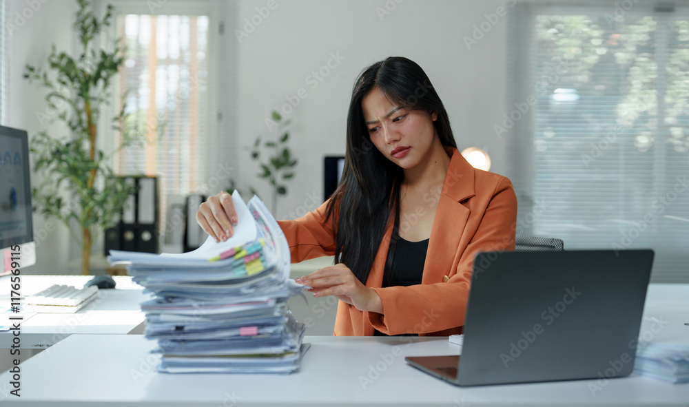 Young, stressed asian businesswoman sifting through a large stack of paperwork while seated at an office desk, surrounded by a laptop and computer, feeling overwhelmed by deadlines Young, stressed asian businesswoman sifting through a large stack of paperwork while seated at an office desk, surrounded by a laptop and computer, feeling overwhelmed by deadlines