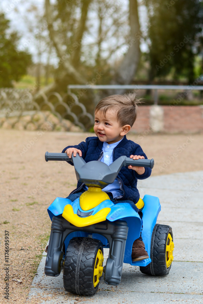 Cheerful toddler riding blue-yellow toy motorcycle, grinning widely amid leafy park setting, experiencing outdoor playtime joy Cheerful toddler riding blue-yellow toy motorcycle, grinning widely amid leafy park setting, experiencing outdoor playtime joy