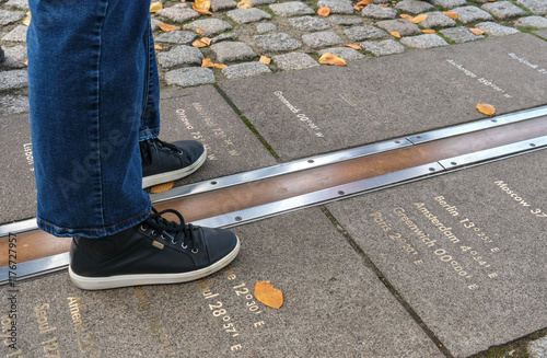 Feet straddling the Prime Meridian at Greenwich, London, with longitude markers engraved on the ground, representing global time zones.