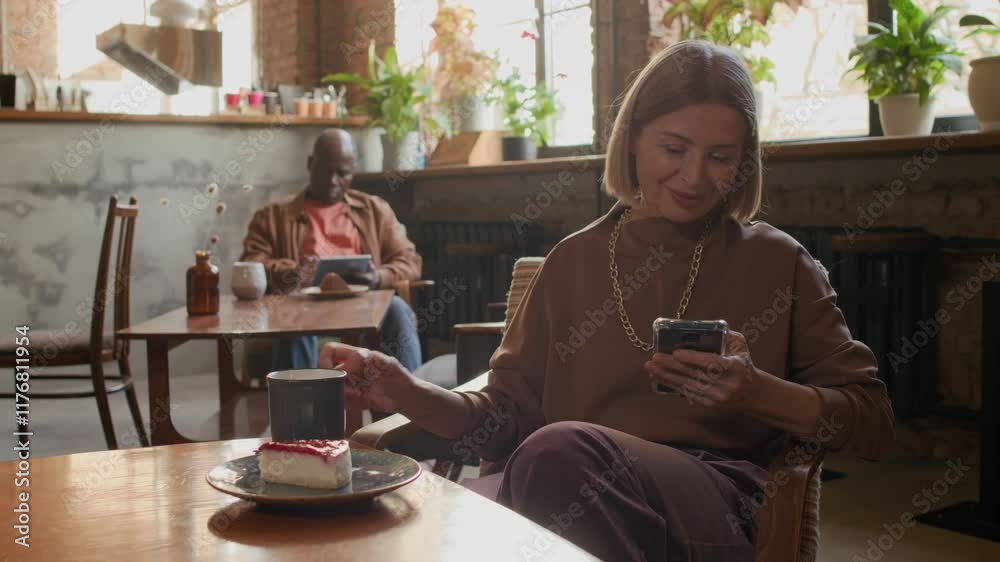 Medium shot of elegant mid-adult Caucasian blonde woman sipping coffee and telecommunicating on smartphone while enjoying her me-time in cozy cafe with rustic design