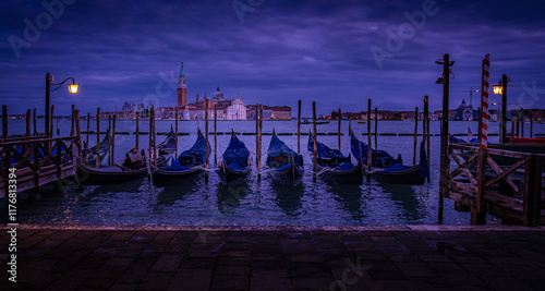 Glowing street lamps illuminate gondolas near serene Venice waterfront during blue hour with Island San Giorgio Maggiore as backdrop