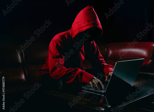 Hackers wear red hoody to cover her faces and sitting in the red sofa dark room. Hacking steal important information using laptop computer to release malware viruses. Ransom and harass organizations.