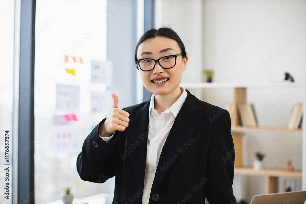 Portrait of young Asian businesswoman showing thumbs up in office, expressing confidence and positivity. Professional female in formal attire standing by window with modern interior.