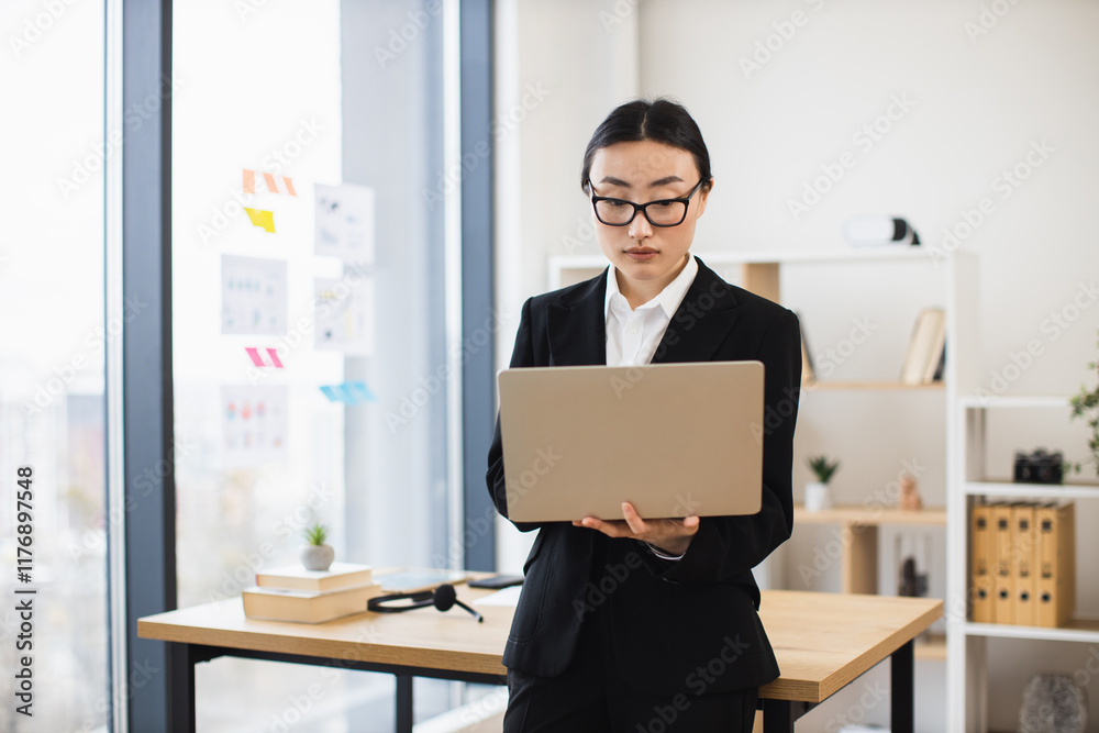 Asian female business professional holding laptop in modern office setting. She appears focused and engaged, standing by desk with large window view, showcasing corporate atmosphere. Asian female business professional holding laptop in modern office setting. She appears focused and engaged, standing by desk with large window view, showcasing corporate atmosphere.