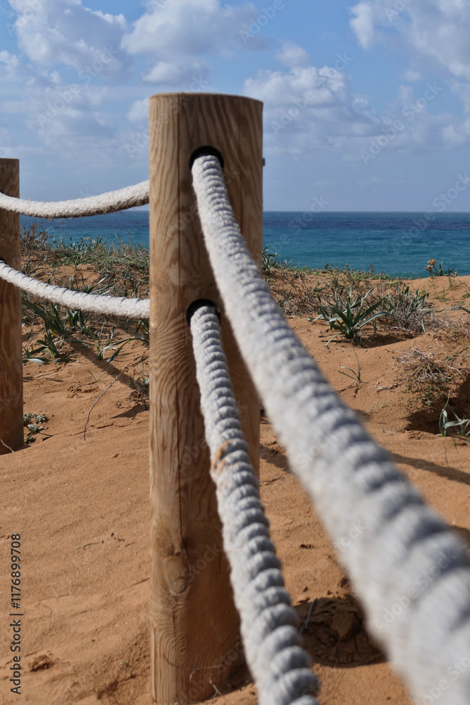 Obraz premium Rope fence on the beach with blue sky and sea in background in Netanya. Sturdy rope fence, composed of thick, grey ropes secured between light brown wooden posts