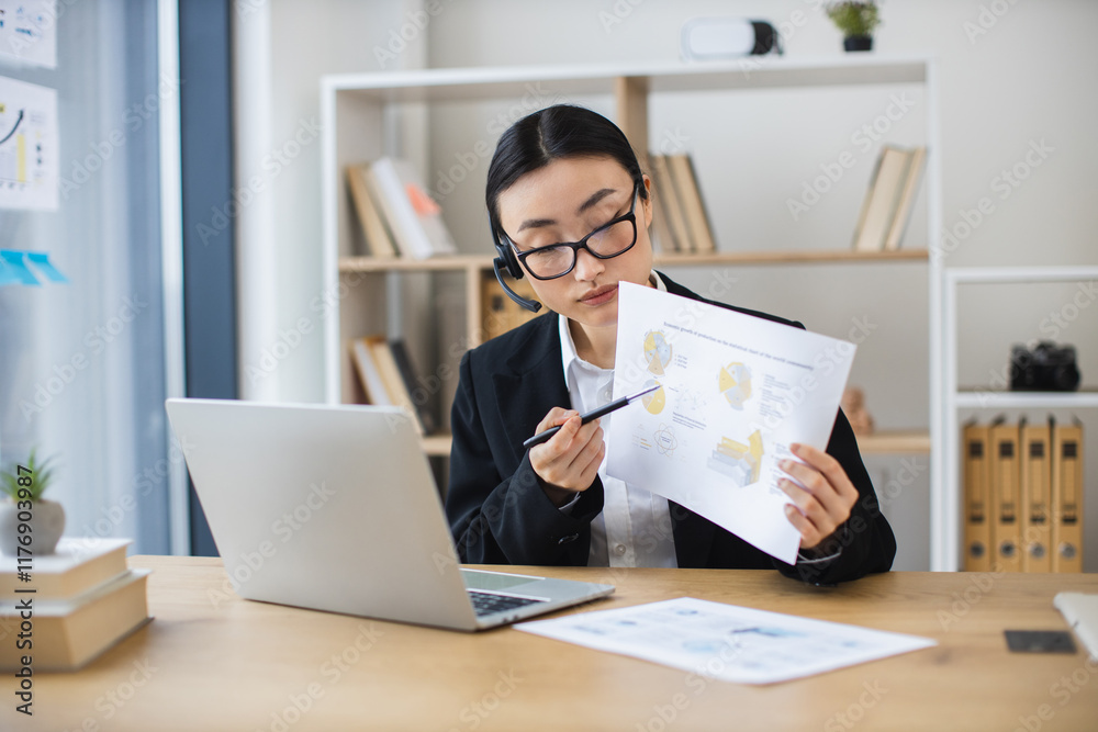 Asian businesswoman wearing headset engages in online video call while reviewing documents at modern office desk. Asian businesswoman wearing headset engages in online video call while reviewing documents at modern office desk.