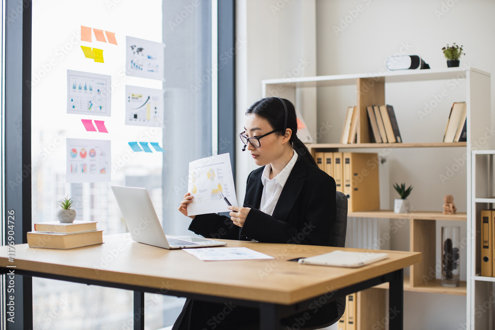 Fototapeta premium Confident Asian businesswoman wearing headset presenting data during video call using laptop. Middle-aged woman working remotely in office setting showcasing professional skills and communication.