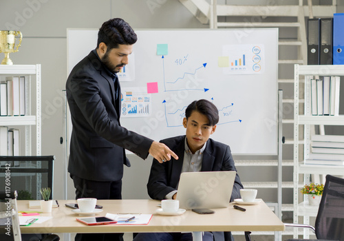 Two professional in suit engage in a discussion at a desk. One point at a laptop screen while the other attentively listen. Chart and graph on the wall provide data insight. photo