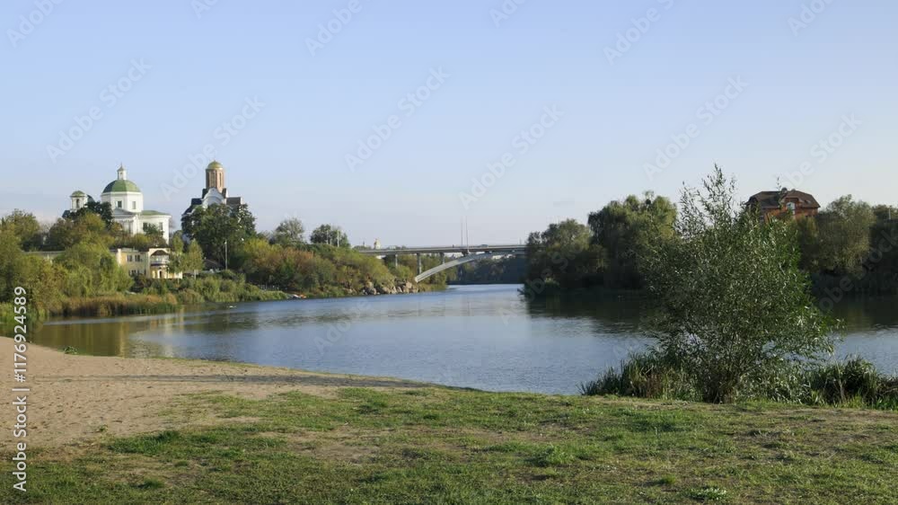 A peaceful riverside scene with lush greenery, a bridge, and historic buildings, including a church with a dome. The calm water reflects the serene atmosphere of the sunny day. A peaceful riverside scene with lush greenery, a bridge, and historic buildings, including a church with a dome. The calm water reflects the serene atmosphere of the sunny day.