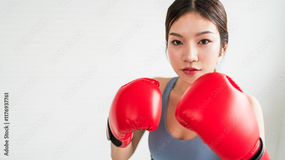 A focused young Asian woman in boxing gloves prepares for a match, showcasing her determination and strength in a fitness studio setting. Indoor working out or exercise at home, wearing sportswear. A focused young Asian woman in boxing gloves prepares for a match, showcasing her determination and strength in a fitness studio setting. Indoor working out or exercise at home, wearing sportswear.