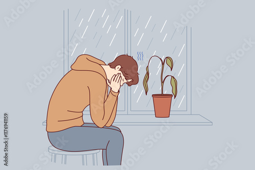 Depressed man sits alone and suffers from melancholy in room with window and wilted flower. Melancholy in older guy victim of autumn blues or everyday problems and troubles at work