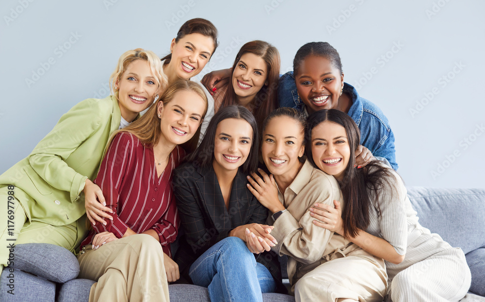 Fototapeta premium Portrait of group of female happy cheerful young diverse women friends coworkers or company employees hugging and looking at camera posing and smiling sitting on sofa on gray background.