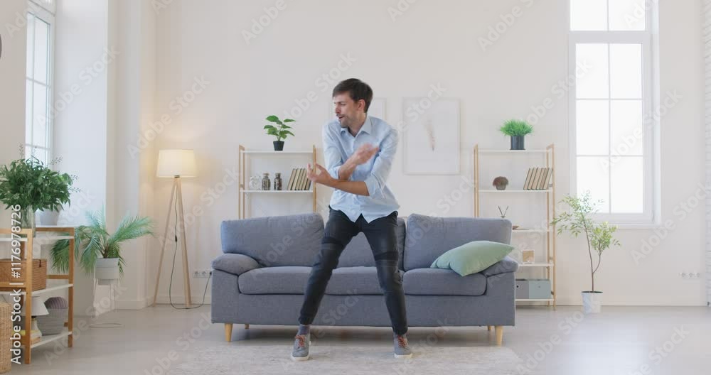 Happy young man in casual clothes having fun during weekend, energetically dancing to music in living room. Cheerful man celebrating success or joyful occasion, fooling around, enjoying free time. Happy young man in casual clothes having fun during weekend, energetically dancing to music in living room. Cheerful man celebrating success or joyful occasion, fooling around, enjoying free time.