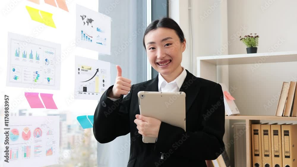 Young Asian businesswoman in office holding tablet giving thumbs up gesture. Positive workplace environment with charts on glass wall. Modern corporate setting promoting teamwork and success. Young Asian businesswoman in office holding tablet giving thumbs up gesture. Positive workplace environment with charts on glass wall. Modern corporate setting promoting teamwork and success.