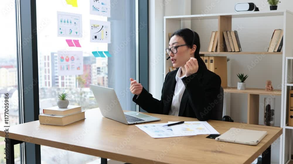 Asian businesswoman in headset conducting video call on laptop in modern office. Woman wearing glasses and suit appears professional and focused, engaging in conversation. Asian businesswoman in headset conducting video call on laptop in modern office. Woman wearing glasses and suit appears professional and focused, engaging in conversation.