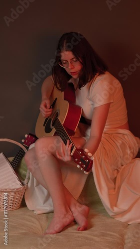Candid female singer songwriter strumming acoustic guitar with enthusiasm, energy and creativity. Musician sitting couch in domestic room bathed in warm, flickering light reminiscent glow of fireplace