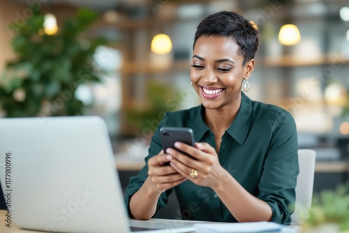 A cheerful woman in a green shirt using a smartphone and laptop at a desk, with blurred office background, representing technology and productivity. Ai generative
