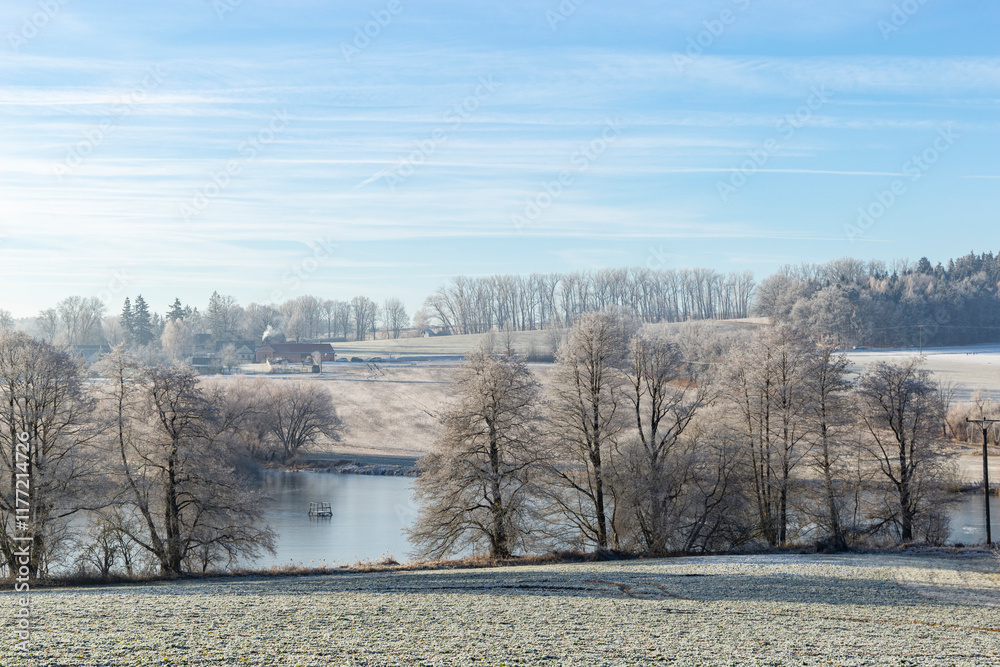 A serene winter morning in the countryside with frost-covered trees, a calm pond, and distant farmhouses under a clear blue sky.