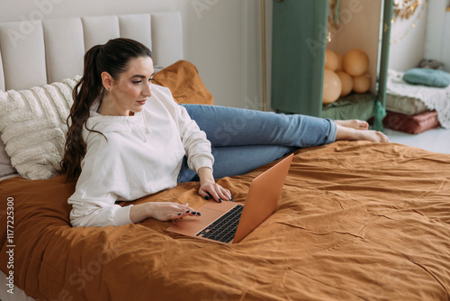 young woman in a hoodie working on a laptop online from home while lying on the bed