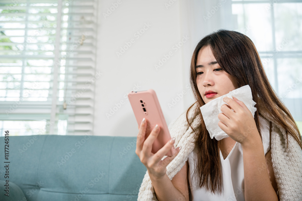 Asian young woman sick sitting down on sofa with cold blowing her nose and checking smart mobile phone to cell to doctor online, depressed sad sick female holding tissues and smartphone in living room Asian young woman sick sitting down on sofa with cold blowing her nose and checking smart mobile phone to cell to doctor online, depressed sad sick female holding tissues and smartphone in living room