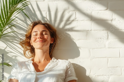 A serene woman relaxes against a white wall, soaking in sunlight and tranquility, surrounded by lush greenery, exemplifying inner peace and contentment amidst vibrant nature.