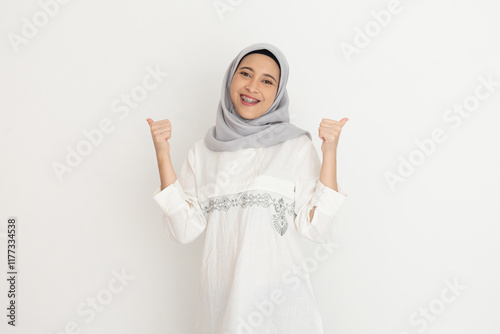 Young muslim woman wearing a grey hijab and white outfit smiles and points to the side while standing against a simple white background presenting something. photo