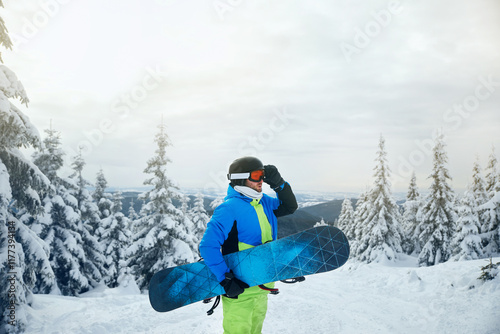 Man with snowboard standing amidst snowy landscape, surrounded by pine trees and a serene background. photo