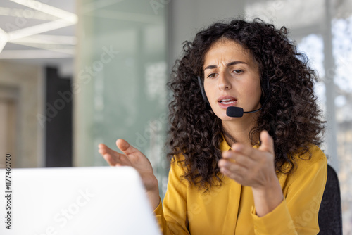 Serious and focused woman with headset talking remotely, close-up of helpline worker working remotely, using laptop for video call, at workplace inside office.