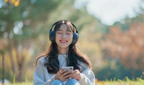 Asian Young Woman Sits in a Park Wearing Headphones and Smiles While Using Her Phone