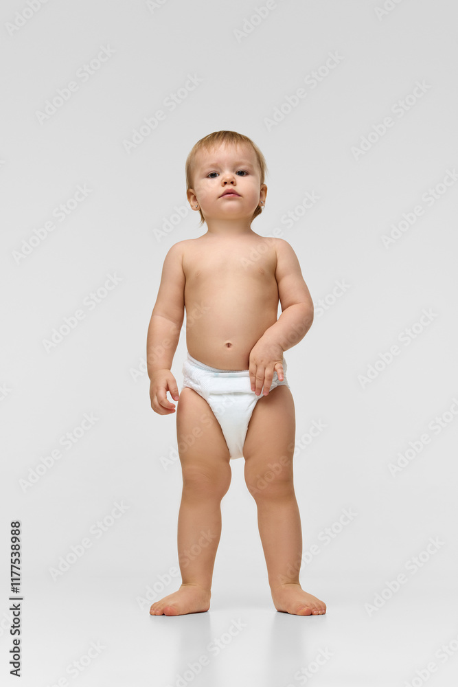 Full-length portrait of little baby girl, toddler standing in diaper with serious face against studio background. Concept of childhood, health, care, parenthood Full-length portrait of little baby girl, toddler standing in diaper with serious face against studio background. Concept of childhood, health, care, parenthood