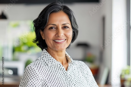 A smiling, middle-aged Indian businesswoman wearing a polka dot shirt, standing in a modern office with ample copy space. Ideal for professional and corporate materials