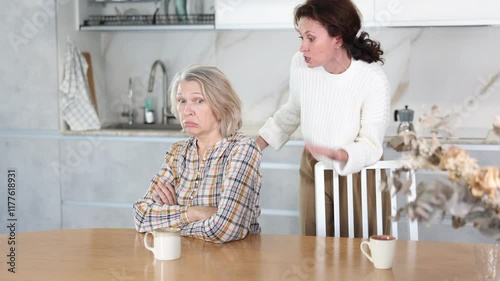 Sad mature woman sitting with a cup in hand and middle-aged female family member brawling to her in the kitchen photo