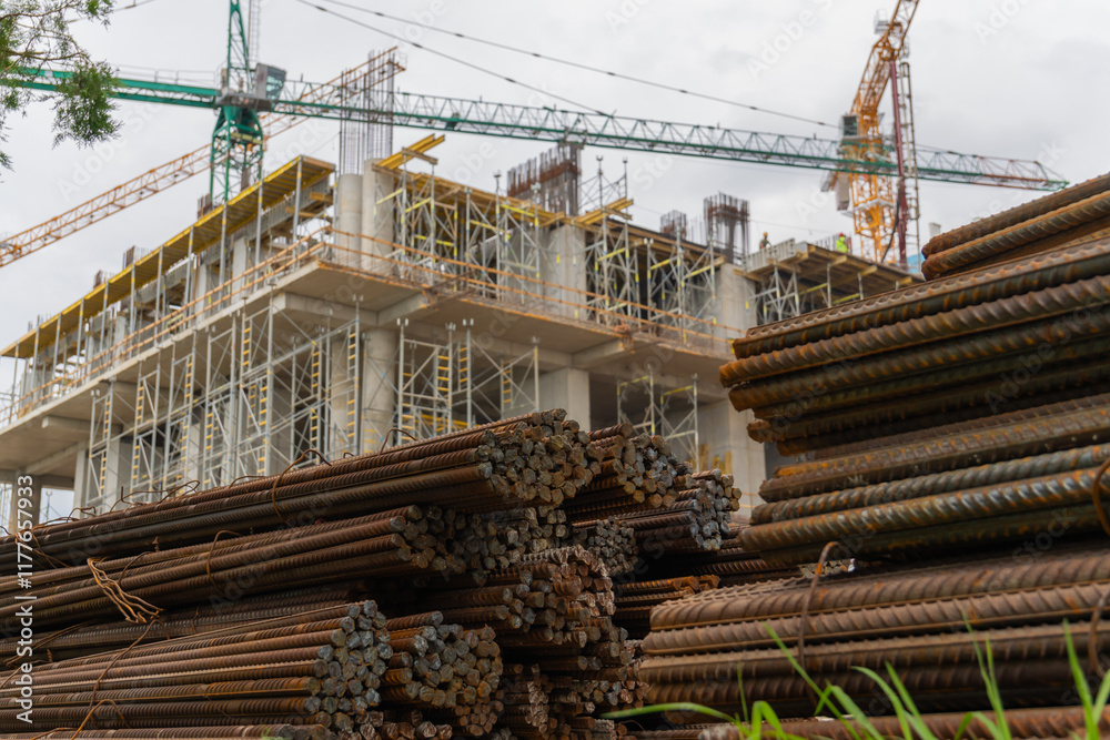 Steel rods stacked together at a construction site, with construction workers and a crane visible working on the building in the background
