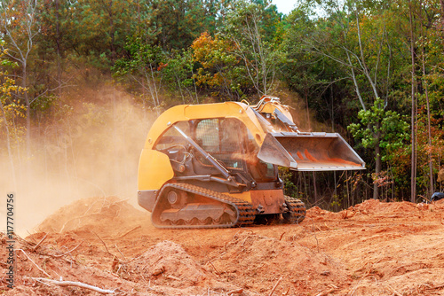 Skid steer loader works on clearing earth amid trees, creating dusty environment during preparing land for development