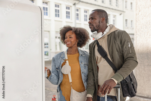 Smiling African American traveler couple buying train tickets at vending machine in a railway station. Travel service and tourism concepts.