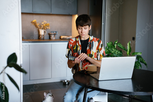 Cat looking up at woman opening pills at table with open laptop photo