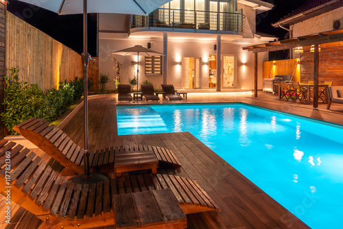 Luxury villa with illuminated swimming pool and wooden loungers at night. Concept of modern backyard relaxation photo